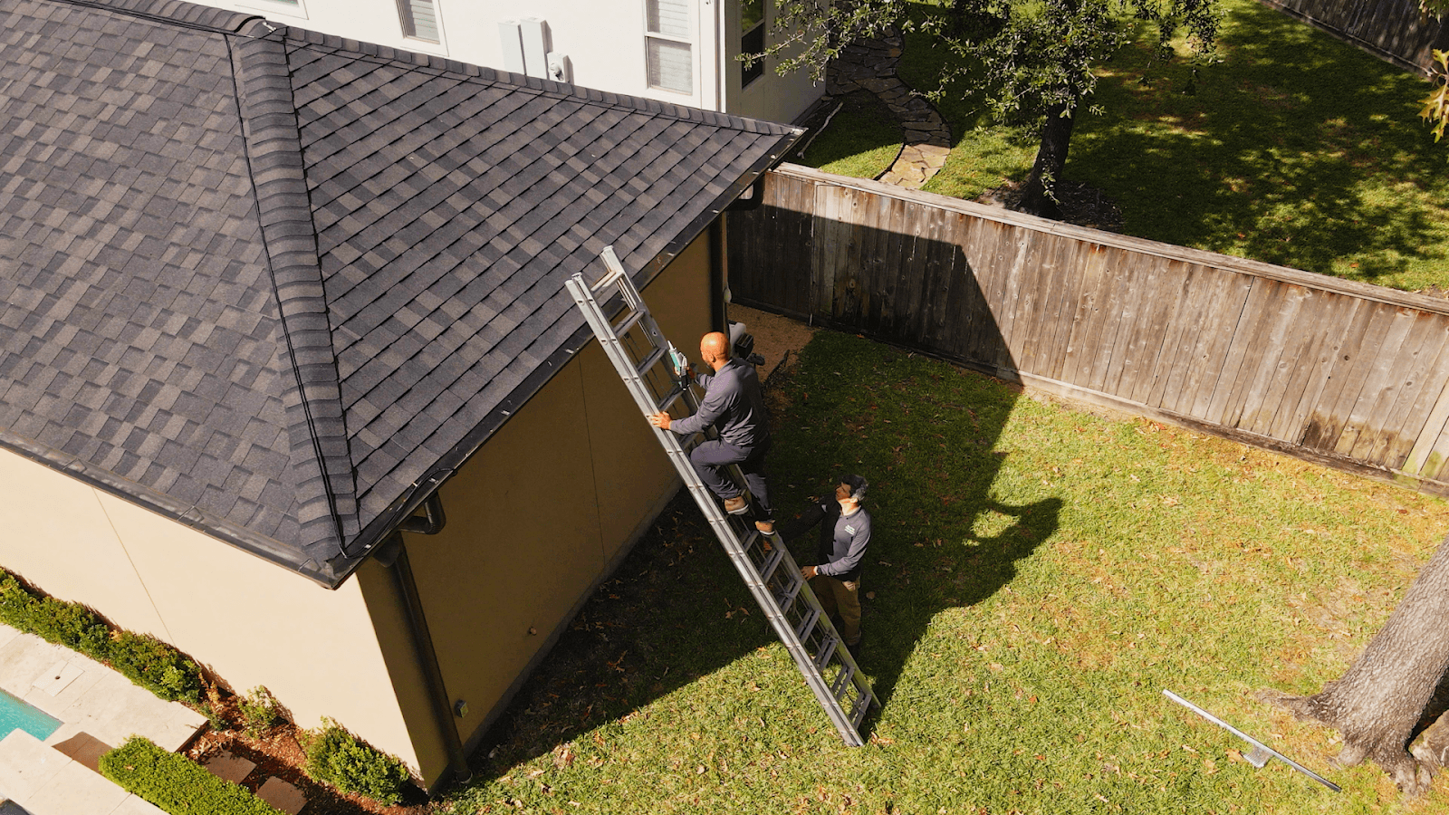 Technician climbing an extension ladder to inspect the roofline of a house while a teammate spots from the lawn