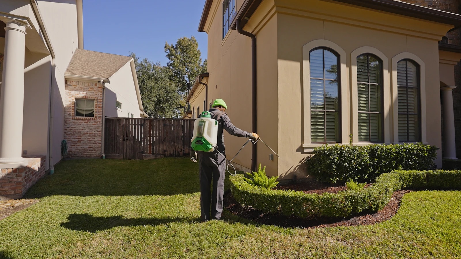 Technician with a backpack sprayer treating the foundation line of a house along a mulched bed